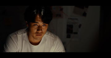 Frame from Heirloom: a man in a white T-shirt lit by a screen in the dark