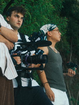 Javi carrying the camera on a support rig beside the assistant camera, foliage behind them