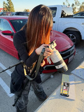 A camera assistant carrying a long lens across a car park